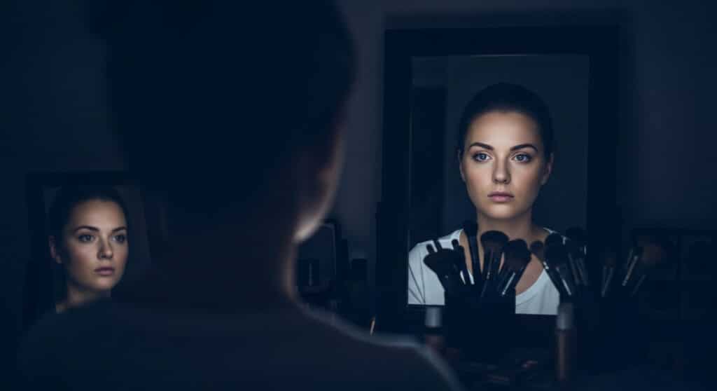 woman staring at her reflection in a dimly lit vanity mirror, surrounded by makeup brushes — a quiet moment showing when beauty backfires