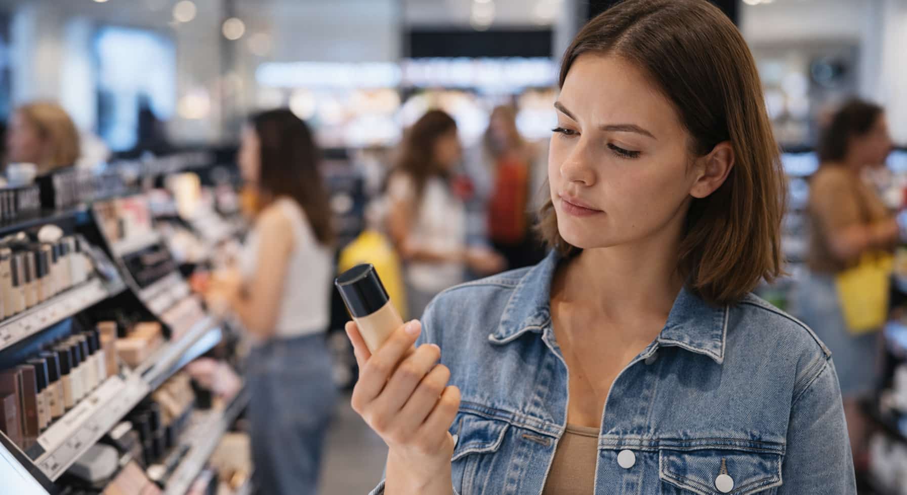 Breaking out from makeup: young woman shopping in a makeup aisle, comparing products while other shoppers browse in the background.