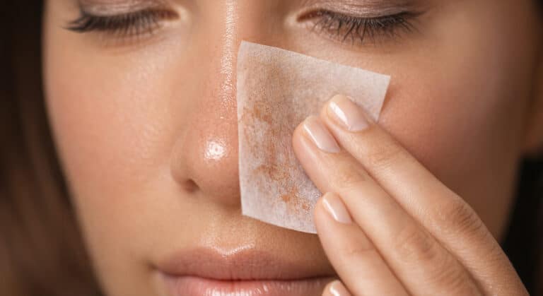 Woman pressing a blotting paper to her nose to absorb excess oil and foundation separation mid-day, visible oil and product residue on the blotting paper.