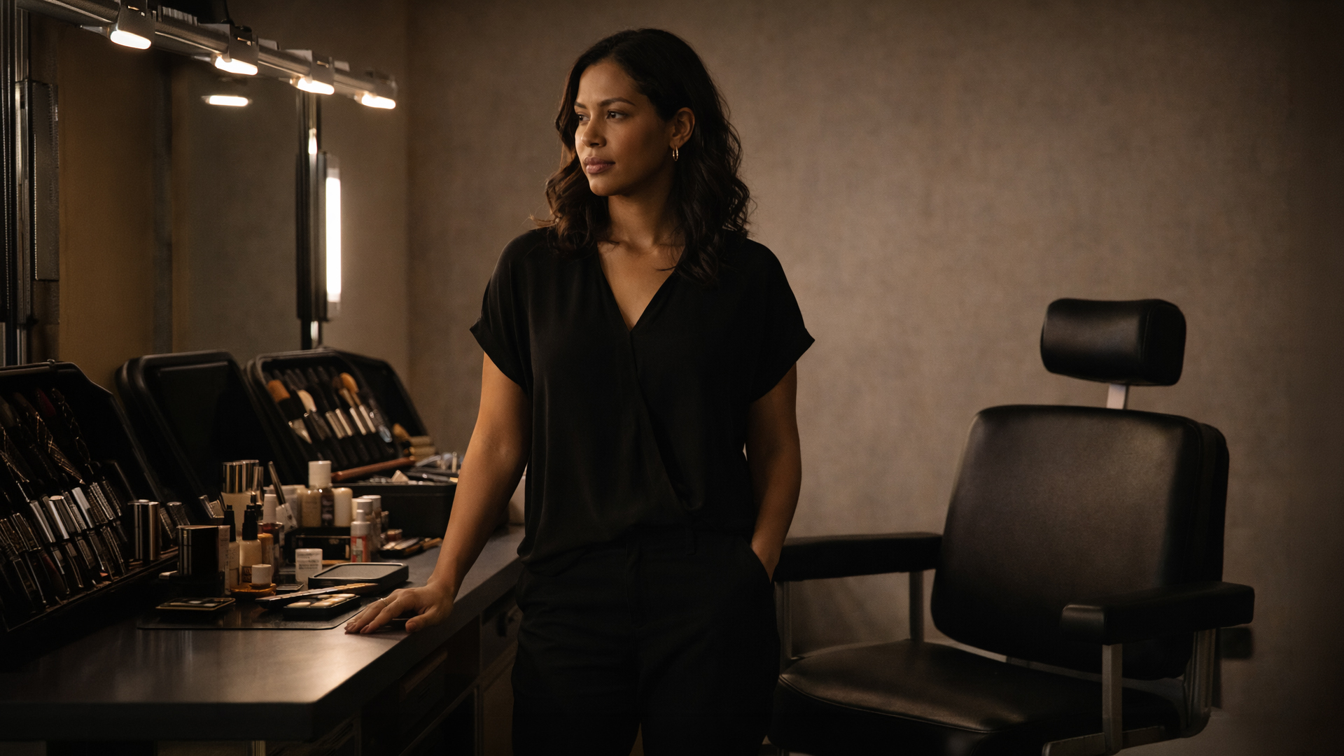 Makeup artist standing at her station beside an empty beauty chair, kit open, expression composed and quietly resolute before a difficult appointment.
